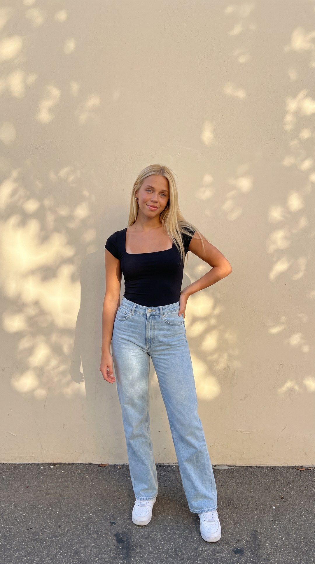 Outfit photo: a woman in a black top and light-wash jeans, hand on hip, against a sunlit cream wall.